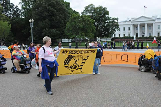 ADAPT members parade in front of the white house.  Two woman carry a giant yellow banner that reads My Medicaid Matters with an ADAPT logo and an outline of the USA.  Tourists  look on from the White House fence.