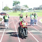 woman pushes man in a wheelchair while other people walk and run behind them on the track. On left side of picture woman walks up to a table.