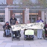 5 of the ADAPT protesters in wheelchairs and standing hold up Not Dead Yet banner