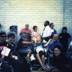 Bryce Thomas and Bob Kafka are surrounded by Texas ADAPTers protesting in front of white brick wall. Also visible are (L-R) Noel Velez, Carolyn Long, and Janet Thomas in foreground