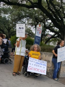 Flanked by two women, Stephanie Thomas in her wheelchair, holds a large sign that reads Denying Disabled People Health Care is Genocide.  