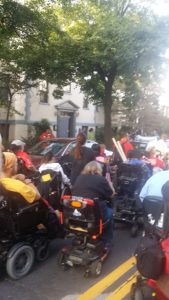 From the back, a group of people in wheelchairs are gathered in the street by parked cars,.  They appear to be heading toward a house. 