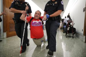 Blind man (Tom Earle) in a red ADAPT T-shirt and holding his white cane in one hand, is dragged down a hallway by two police officers.  An officer on each side holds his arms and his legs drag behind him.