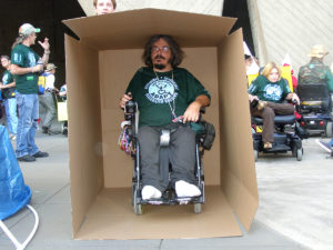 David Wittie sits in his wheelchair in a huge cardboard box. He is wearing his accessible, affordable, integrated housing ADAPT shirt and behind the box you can see other ADAPT members at the national HUD plaza.