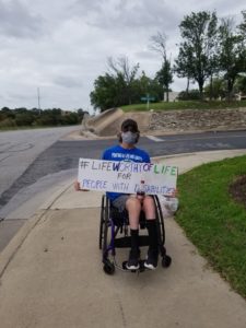 Jeff, sitting in manual wheelchair (and in mask) by side of road holds long sign reading "#Life Worthy of Life for People with Disabilities"