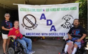 A woman stands next to 2 men in wheelchairs in either side of a banner. It reads Access is a civil right, Our homes not nursing homes ADA Americans with Disabilities Act. The two ADAPT logos (no steps, we will ride and Free our People wheelchair person breaking chains overhead. 