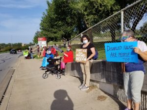 A line of protesters stand on a sidewalk by a chainlink fence. Some are standing, some in wheelchairs.  They are holding posters