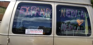 Rear window of van with colorful writing that says Expand Medicaid and Medicare. Below is a bumper sticker that reads Honor thy mother, father, grandma, grandpa keep them out of nursing homes and the o of homes is the ADAPT free our people logo
