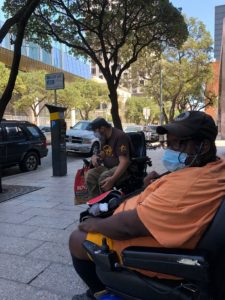 Two men in wheelchairs, one white and one black, wearing face masks, sit on the sidewalk