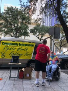 A man with one arm in a red shirt talks with a guy in a tie dye shirt in a wheelchair.  They are in front of van with a Community Integration banner on its side. 