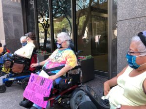 Four people in masks and in their wheelchairs sit in front of plate glass windows of an office building.