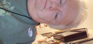 Woman with short white hair lifts her shoulder to show her I voted sticker on her shirt.