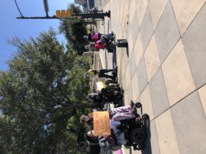 people in wheelchairs and folks standing gather on sidewalk. Trees behind them.