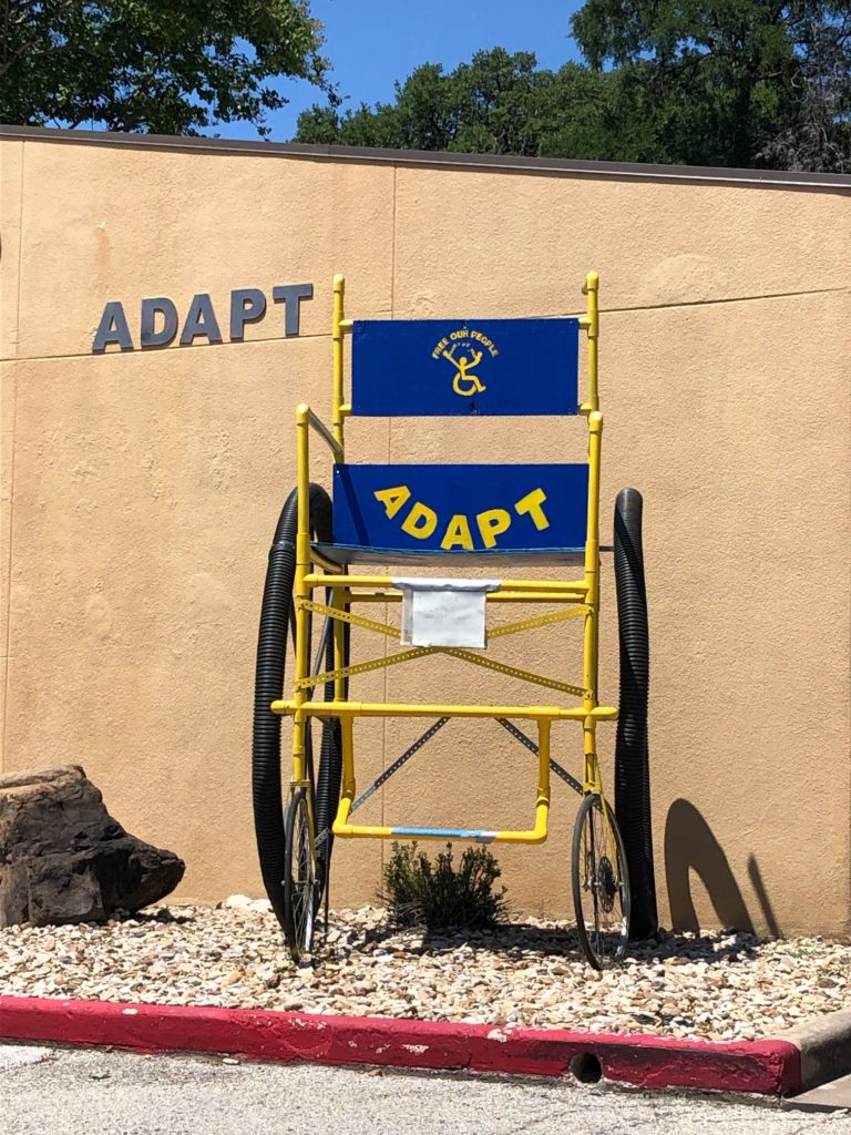 10 ft tall wheelchair painted blue and yellow with the ADAPT Free Our People logo on the back of the seat.  It's in front of the ADAPT of TX office.