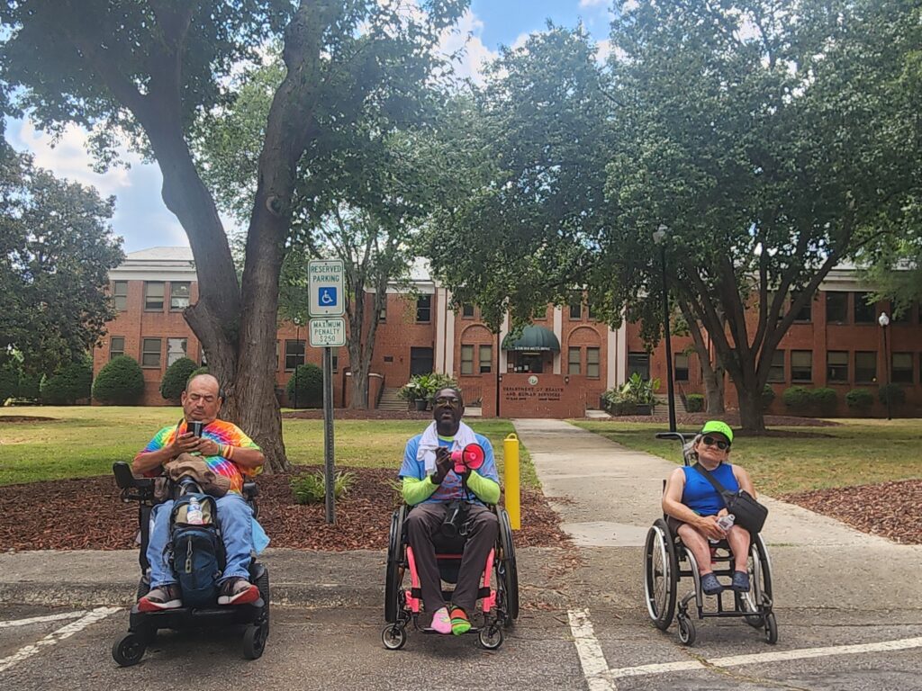 Three ADAPTers in wheelchairs lined up in front of NC HHS building.