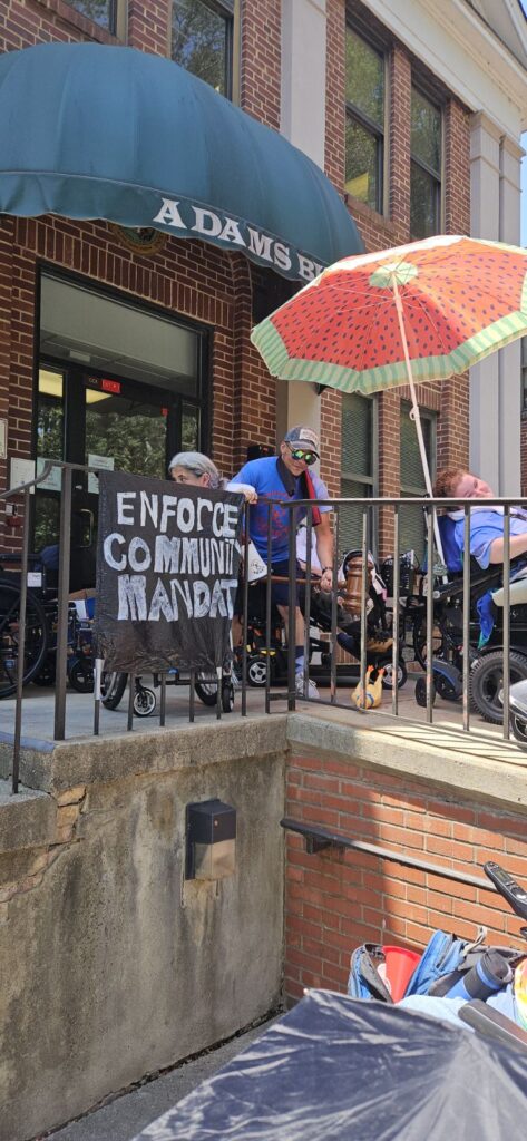 ADAPTers with banner, huge gavel and rubber chicken fill tiny porch in front of HHS state offices.